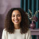 A smiling woman, late 20s, diverse, looks at the camera. The background is blurred with hints of deep plum and dusty lilac.